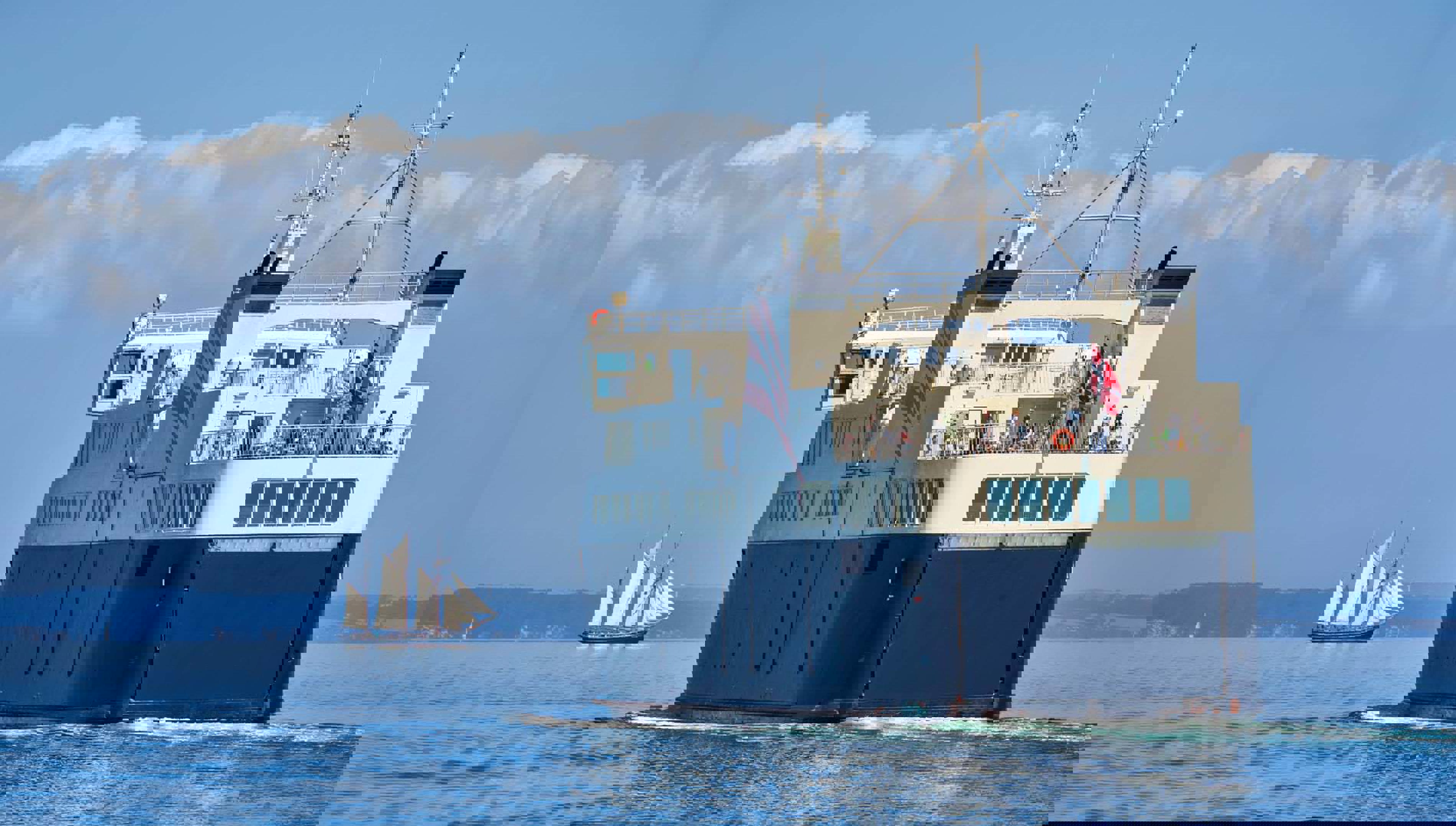 Alslinjen ses sejle fra havn, hvor man i horisontent kan se land of et sejlskib.