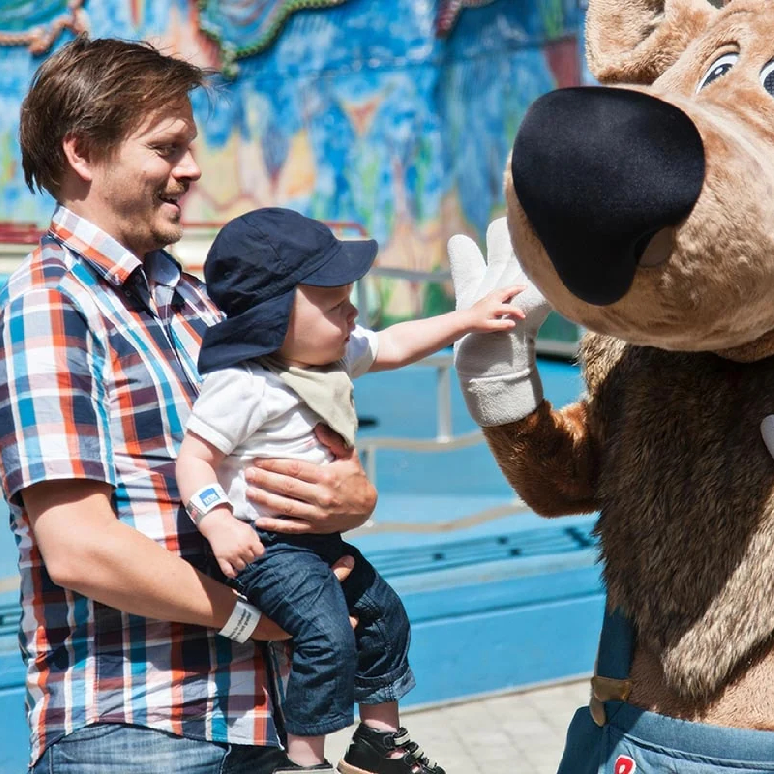 En man i rutig skjorta håller ett barn som rör vid en maskot i en nöjespark.