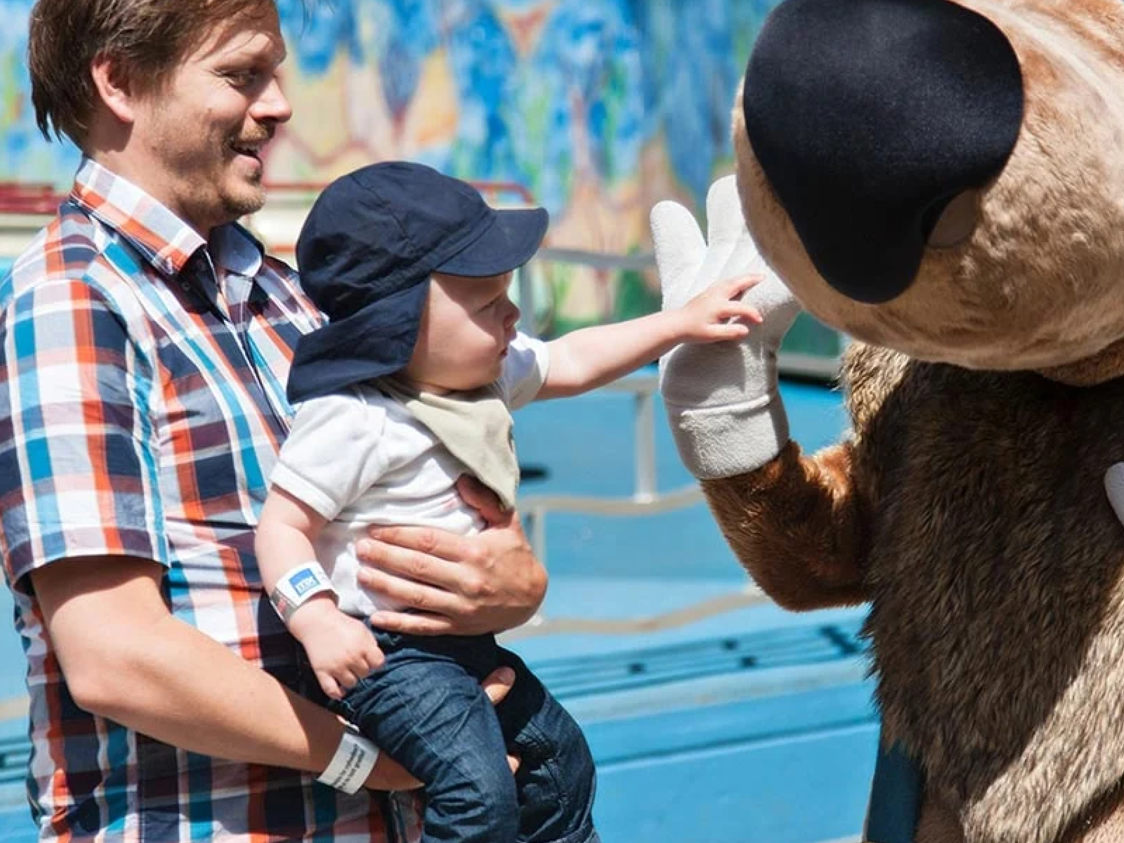 En man i rutig skjorta håller ett barn som rör vid en maskot i en nöjespark.