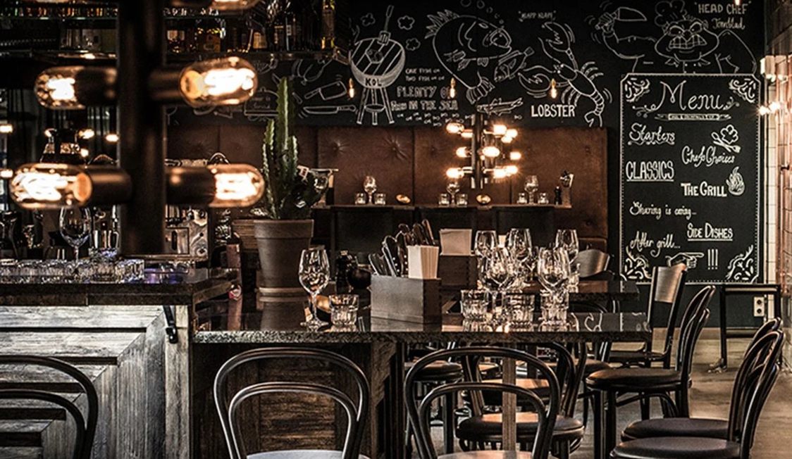 Restaurantinteriør med elegant bordopsætning og sorte bistrostole foran dekoreret kridttavlevæg.