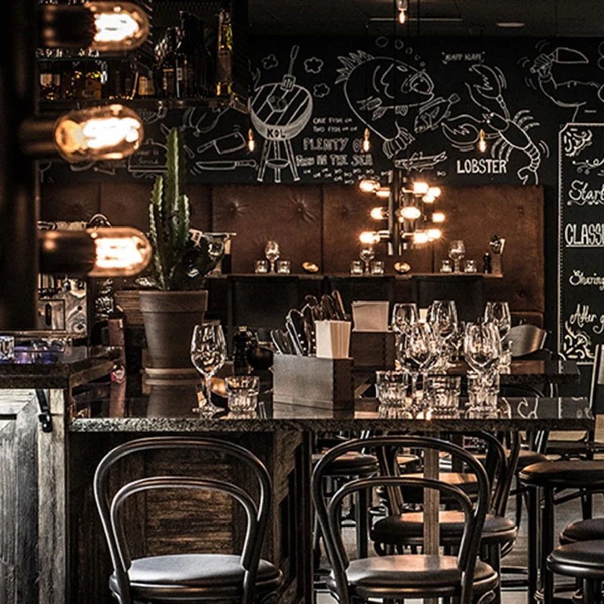 Restaurantinteriør med elegant bordopsætning og sorte bistrostole foran dekoreret kridttavlevæg.