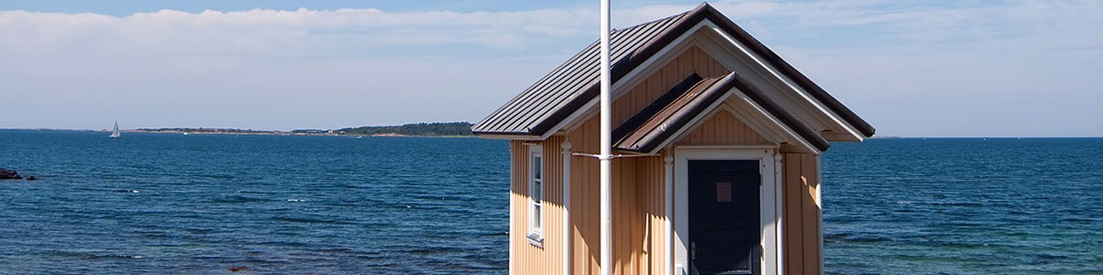 Lille strandhus i træ med sort tag og ensom flagstang ved kystlinje med blåt hav og himmel i baggrunden.