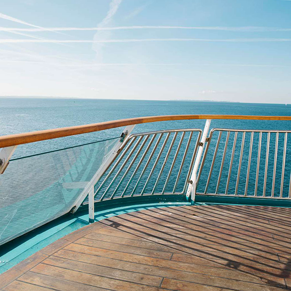 Sjöutsikt från fartygets däck med modernt glasräcke och trägolv under en klarblå himmel.