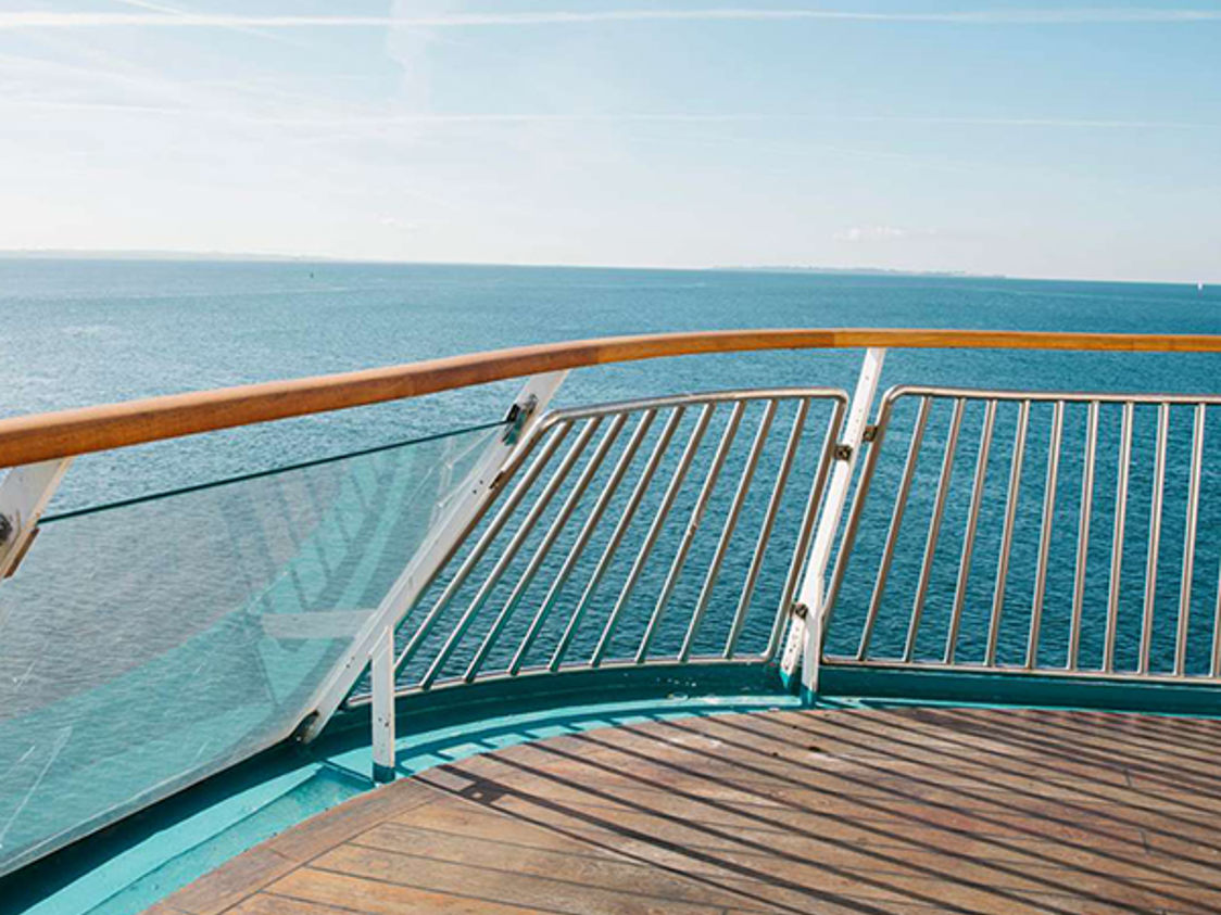 Sjöutsikt från fartygets däck med modernt glasräcke och trägolv under en klarblå himmel.