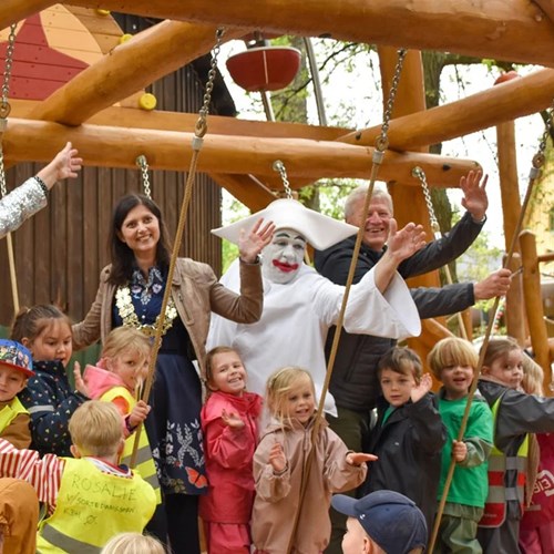 Klovn i hvidt kostume omgivet af glade børn og voksne på en legeplads, der alle vinker til kameraet.