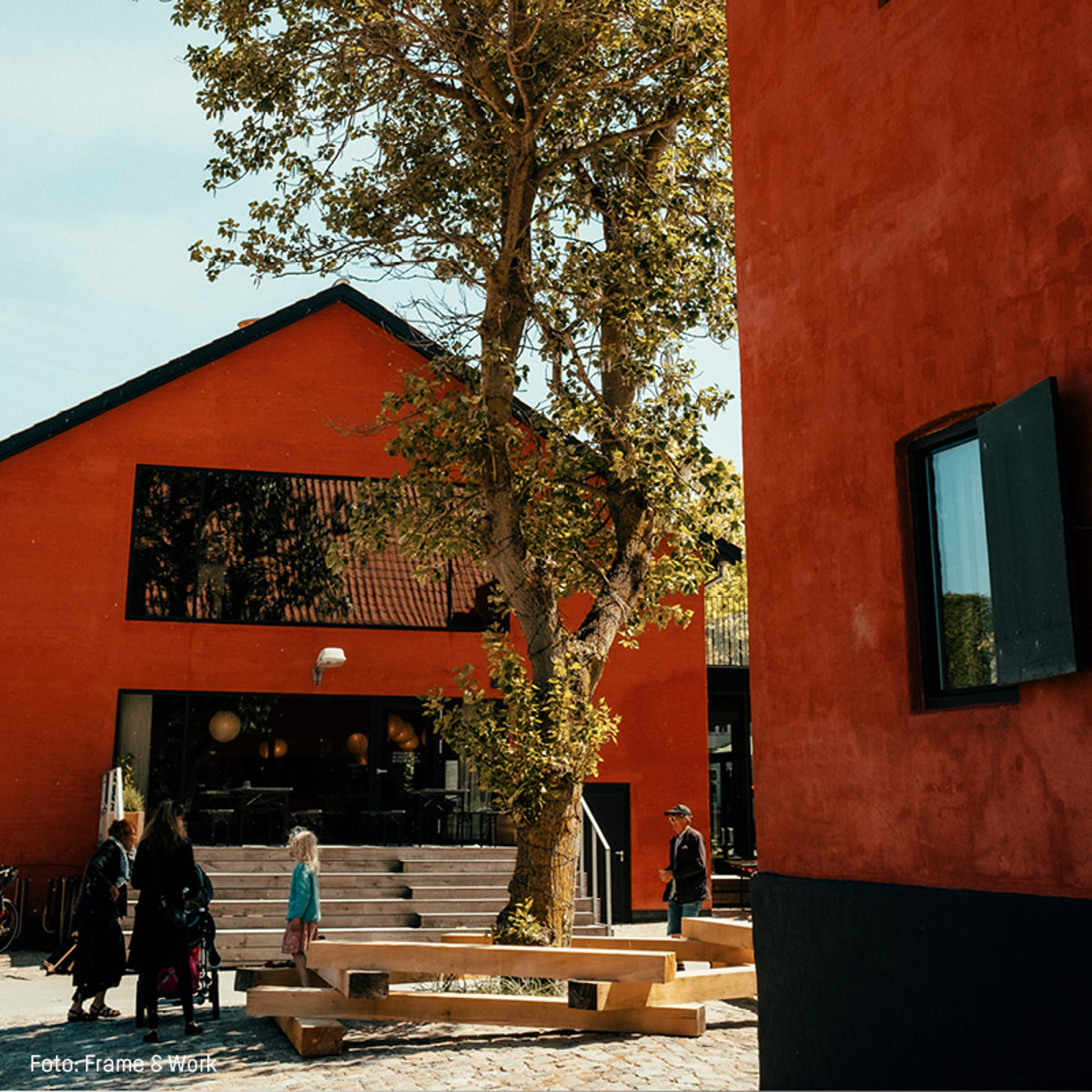 Moderne rød træbygning med glasfacade, træer og besøgende i brostensbelagt gårdhave i solskin.