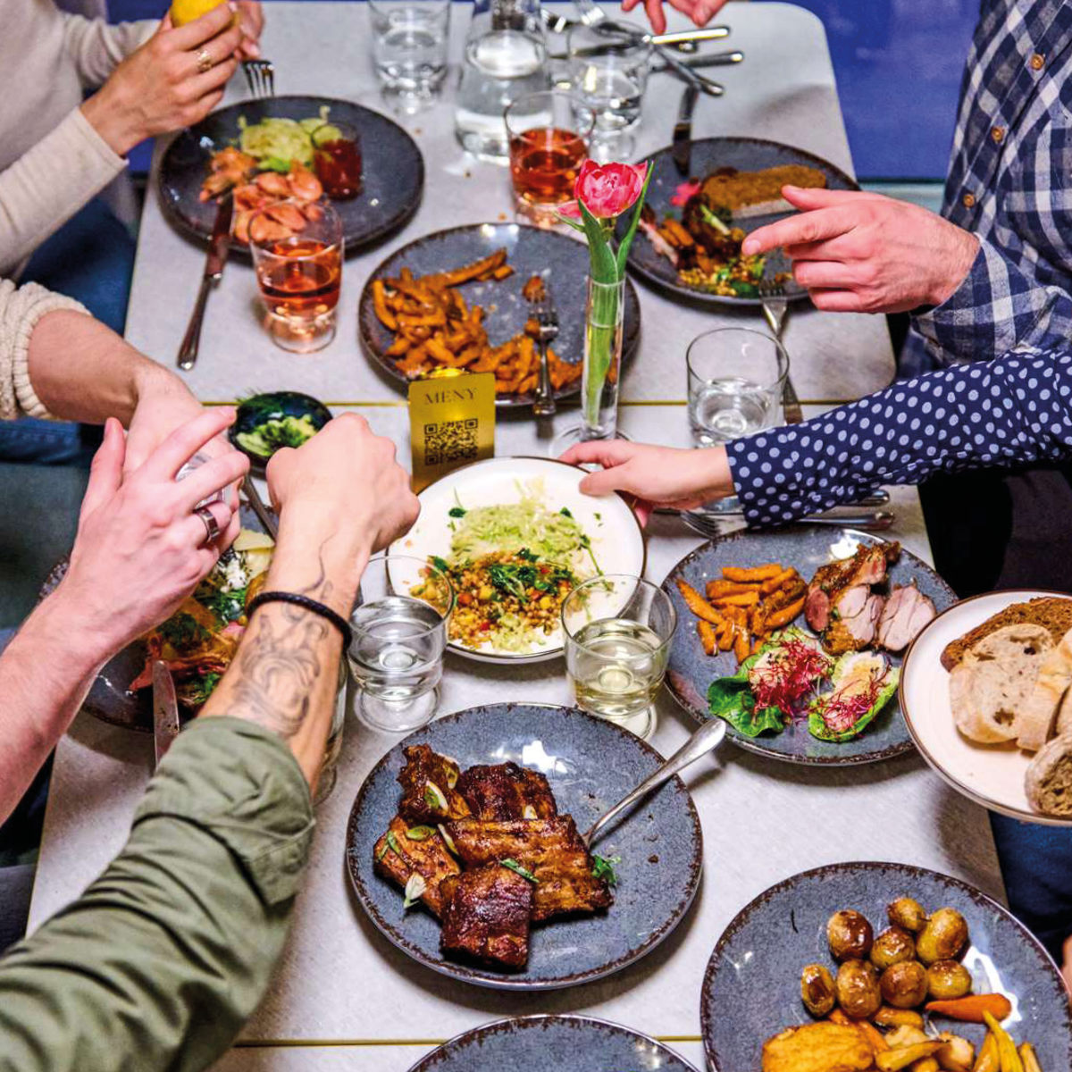 Spisende i restaurant nyder fælles middag med grillmad, salater og stegte kartofler på et bord set i fugleperspektiv.