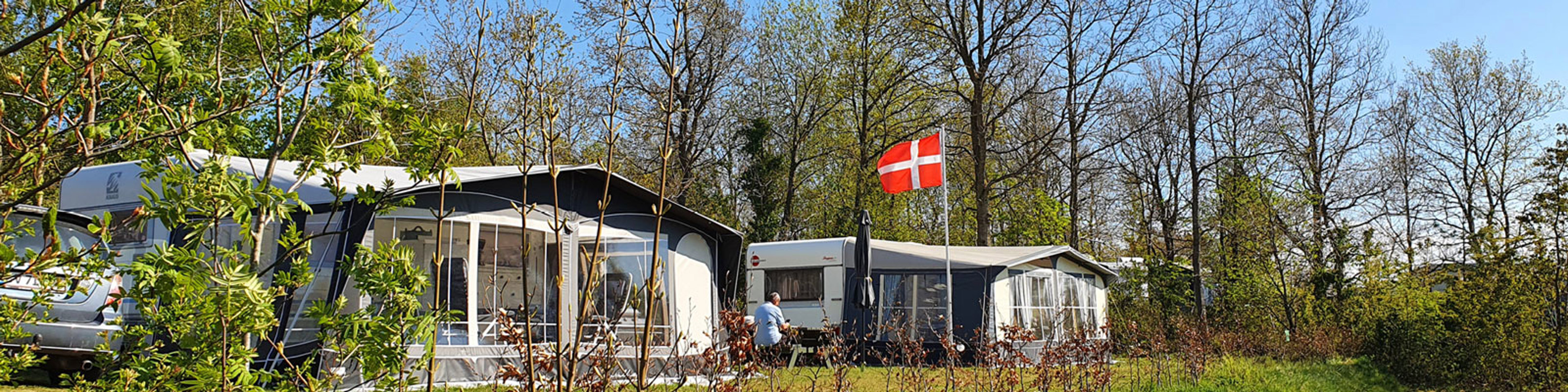 Campingvogn med fortelt i en grøn campingplads omkranset af træer og dansk flag blafrende i vinden.