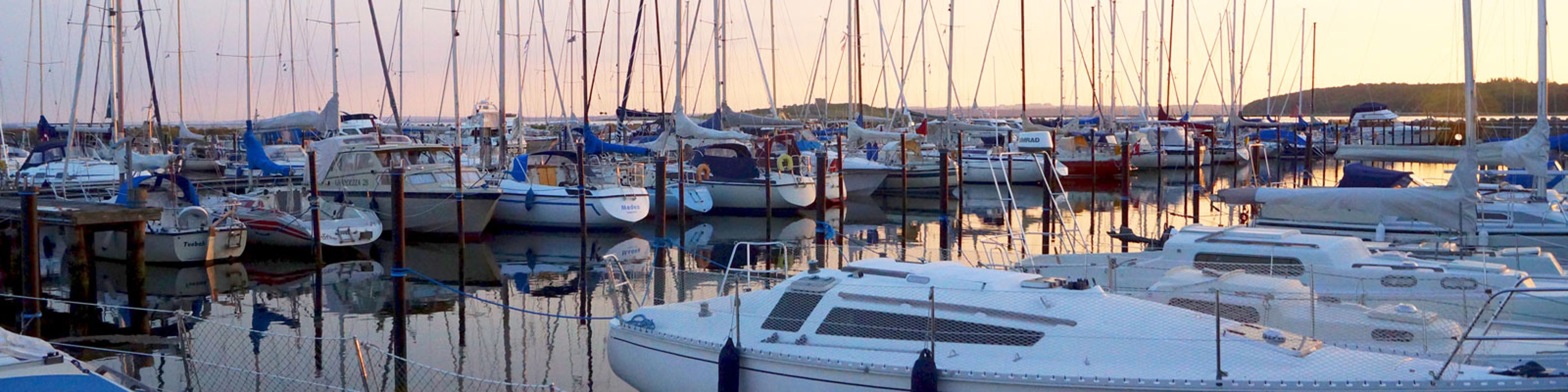 Sejlbåde fortøjet i en marina ved solnedgang, spejler sig i det rolige vand og skaber en fredfyldt atmosfære.