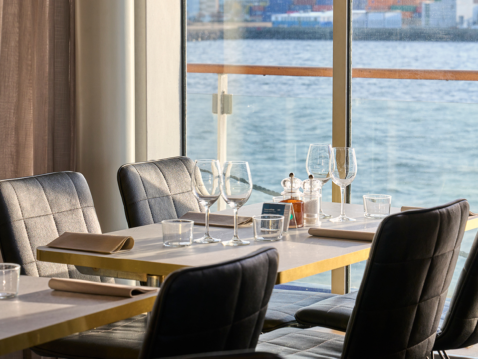 Moderna restaurangbord och stolar vid fönster med havsutsikt, dukade för gäster med vinglas och bestick.