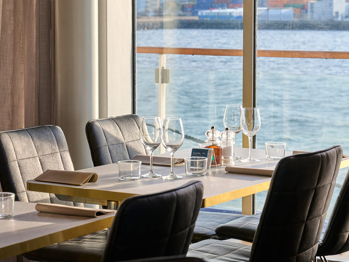 Moderna restaurangbord och stolar vid fönster med havsutsikt, dukade för gäster med vinglas och bestick.