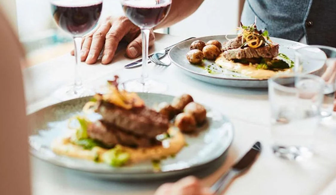Bøf serveret med kartofler og sauce på restaurantbord med to glas rødvin og to personer i fokus.