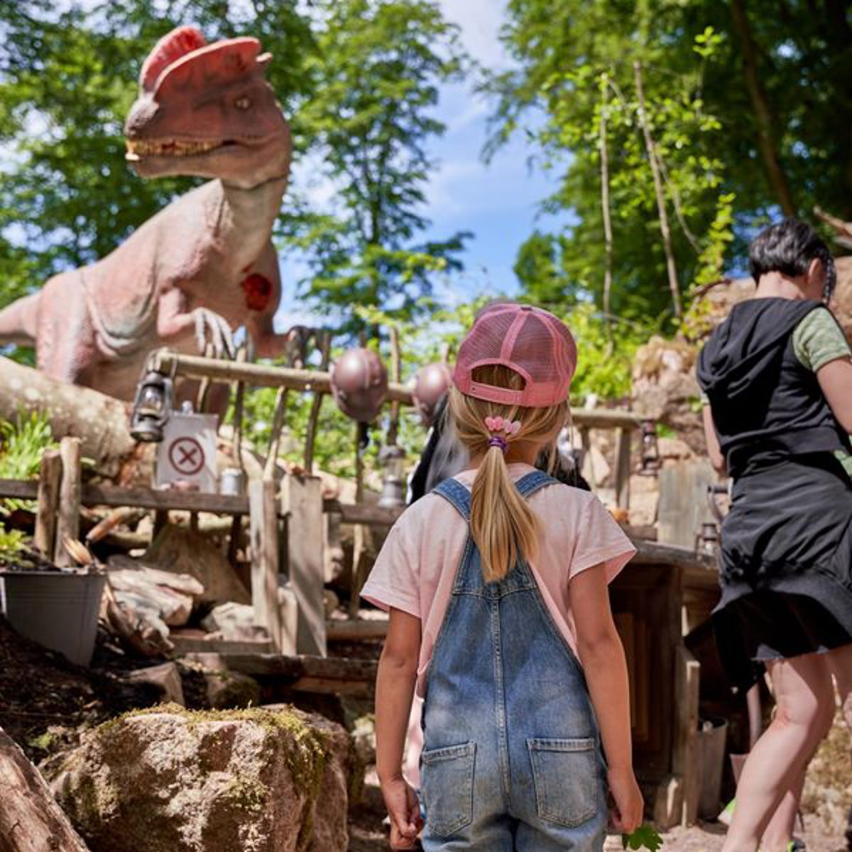 Pige i lyserød kasket kigger på stor dinosaurskulptur i skovområde ved attraktion.