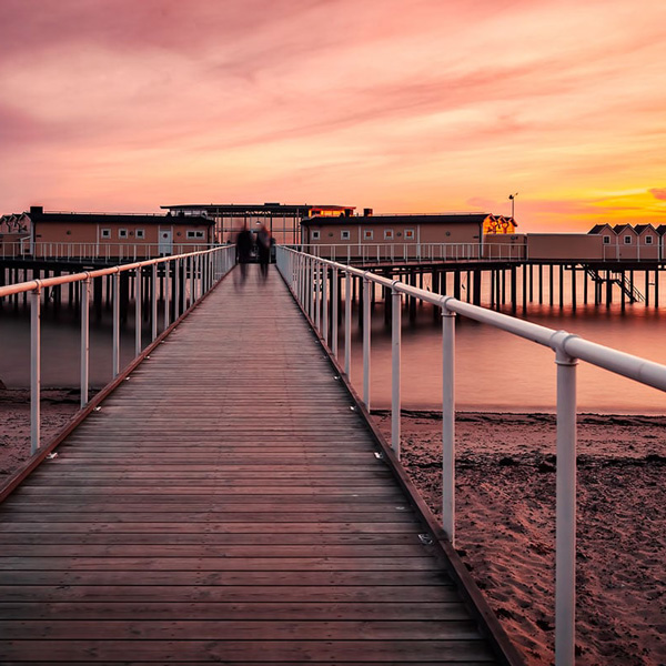 Lang gangbro mod historisk badehus i svenske Landskrona ved solnedgang, set mod horisonten.
