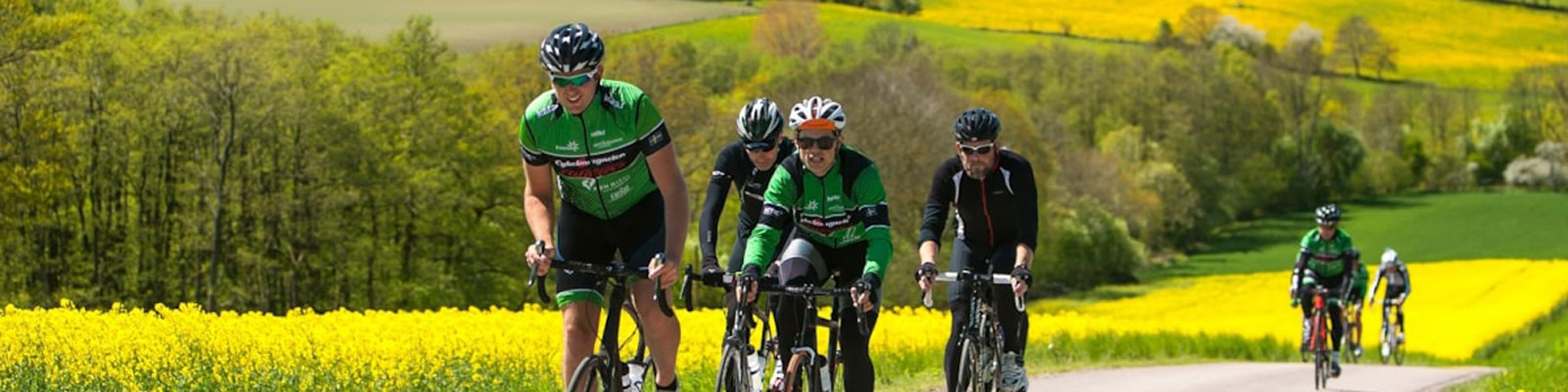 Cykelryttere i grønne trøjer kører sammen på en landevej omgivet af blomstrende rapsmarker i foråret.