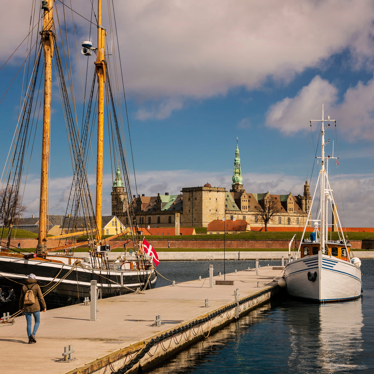 Hamnen med segelbåt och gångbrygga, med utsikt mot Kronborg slott under molnig himmel.