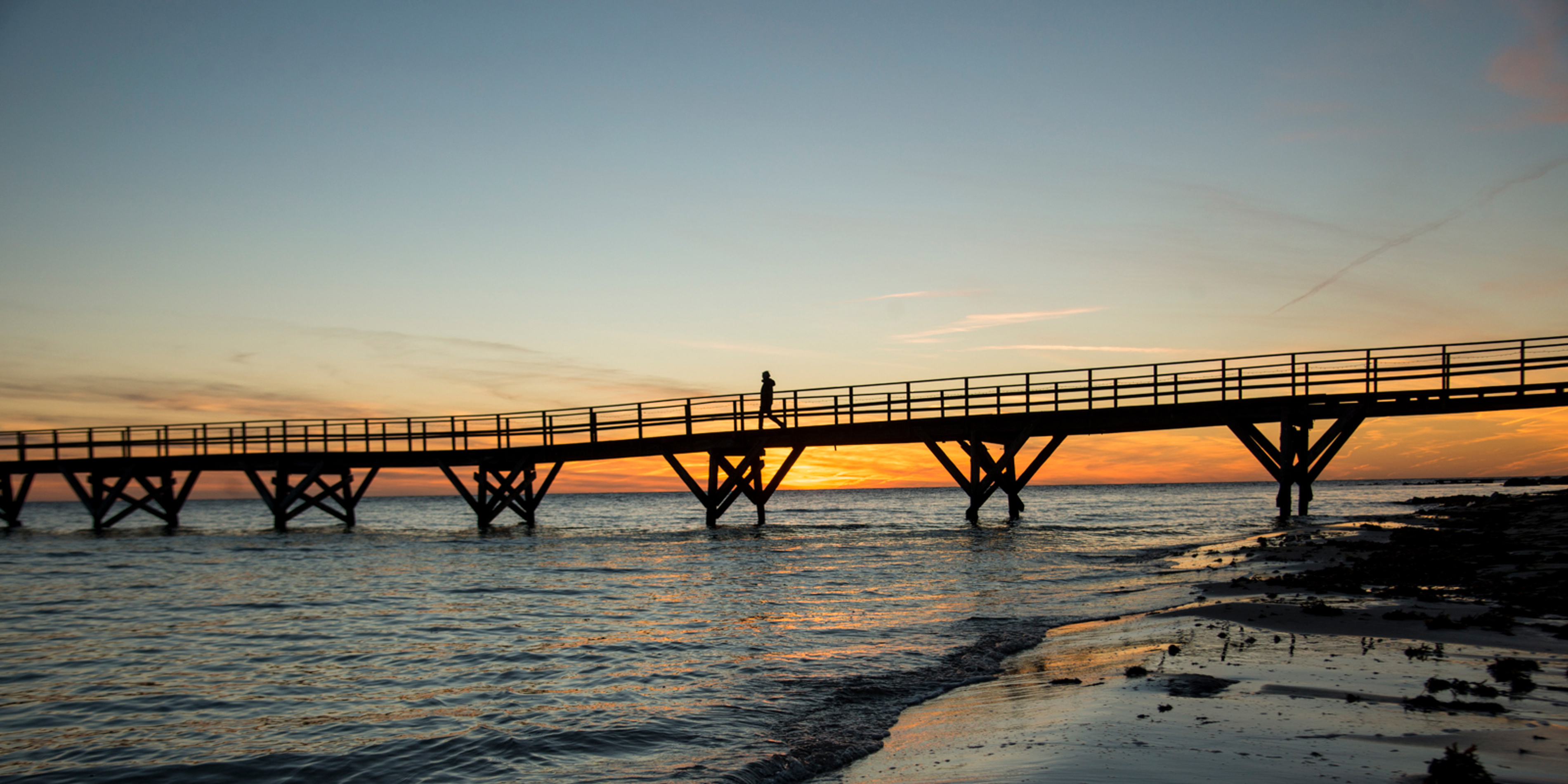 En bro syns i havet med en orange och röd solnedgång i bakgrunden.