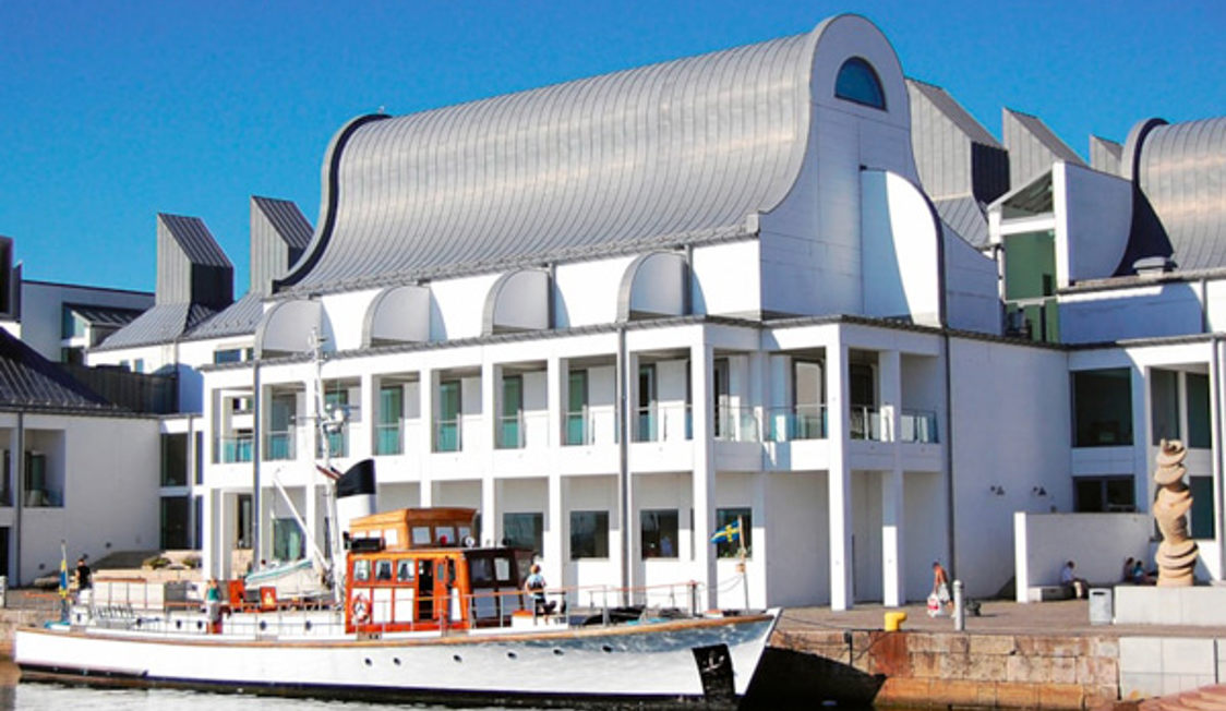 Hvidt, moderne museum med svungne taglinjer set fra vandkanten med en træbåd liggende i havnen foran.