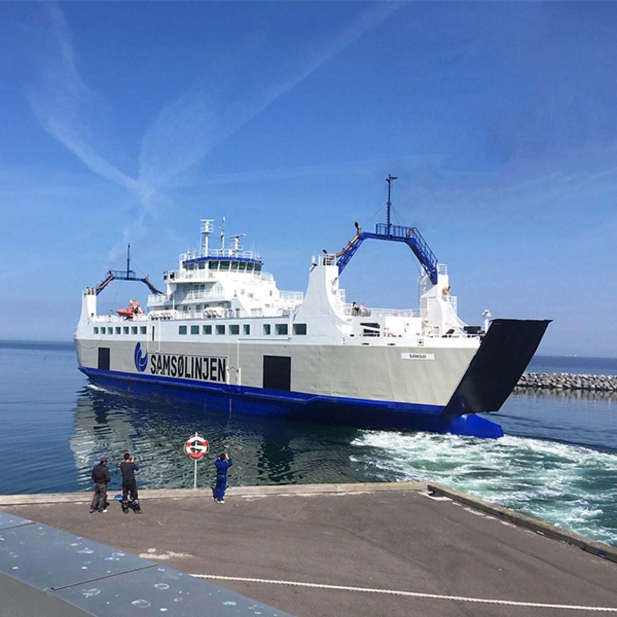 Samsølinjen færge sejler tæt på kaj under blå himmel, set fra havnefronten i en dynamisk vinkel.