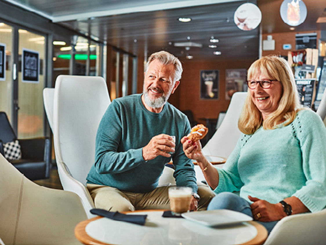 Ett äldre par njuter av en fika med kaffe och bakelse på ett café med moderna stolar.