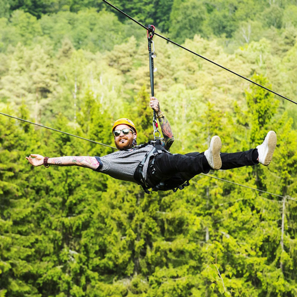 Mand klædt i sort tøj glider i en sele på en svævebane iført gul hjelm over frodig skov.