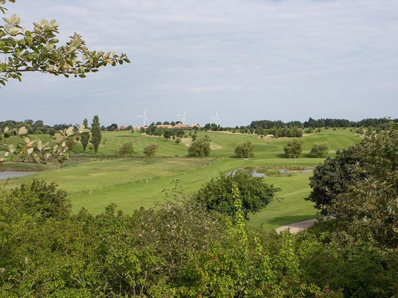 Mon Golf Center med vidsträckt vy av frodig grönska och vindkraftverk i bakgrunden under en molnig himmel.