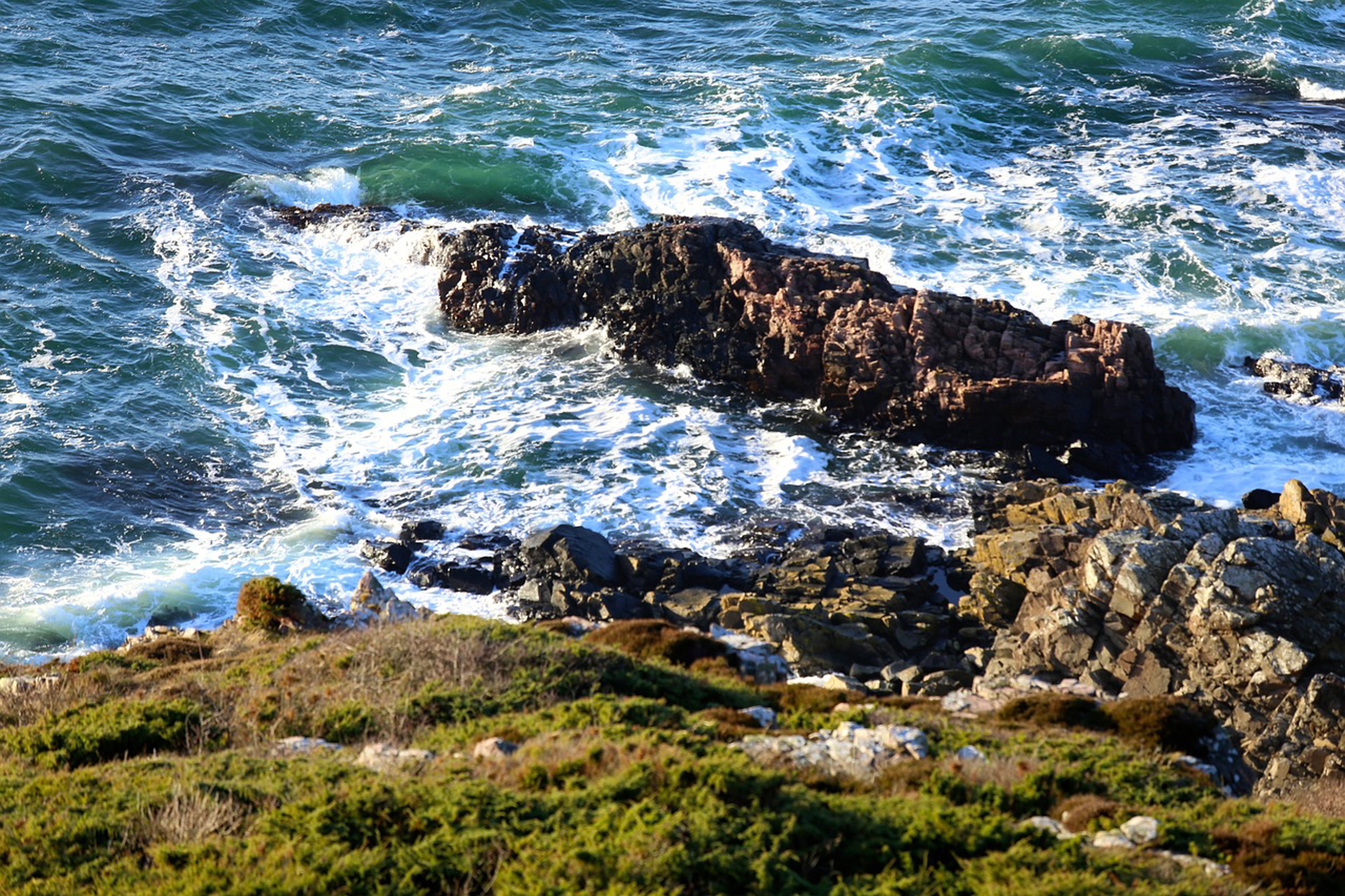 Stenet kystlinje ved havet med bølger, der slår mod klipperne, set fra toppen af en bakke.