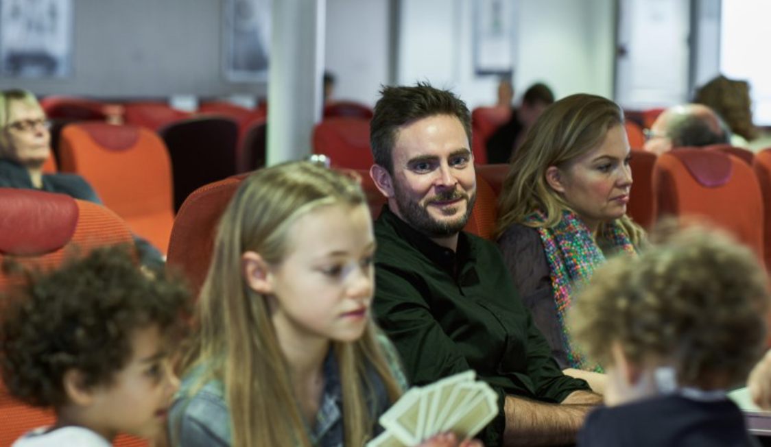 Man ler medan han sitter i en färjesätebukt, omgiven av andra passagerare, med barn som spelar kort framför.