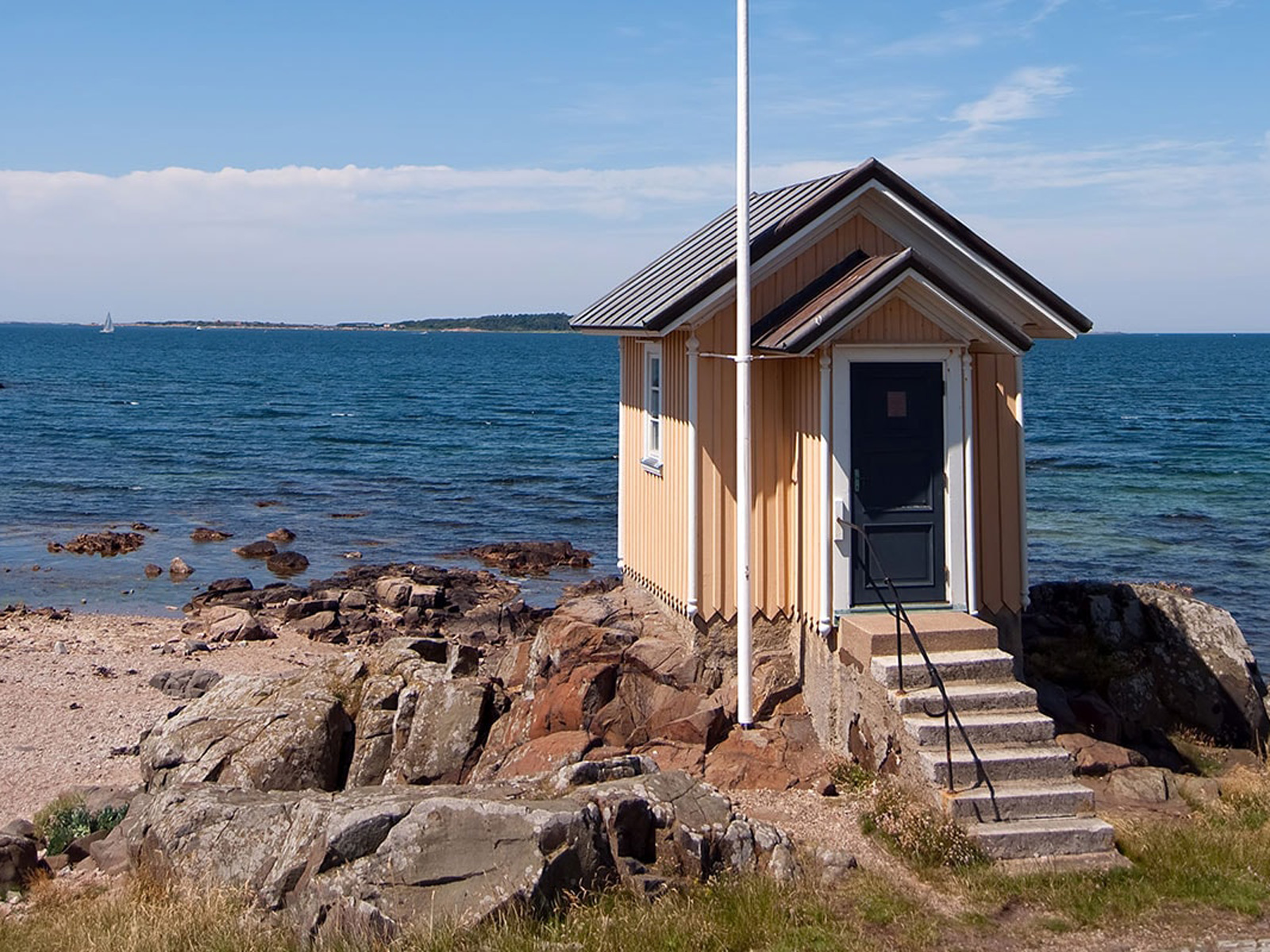 Lille træhus i gule nuancer stående på klipper ved havet under blå himmel.