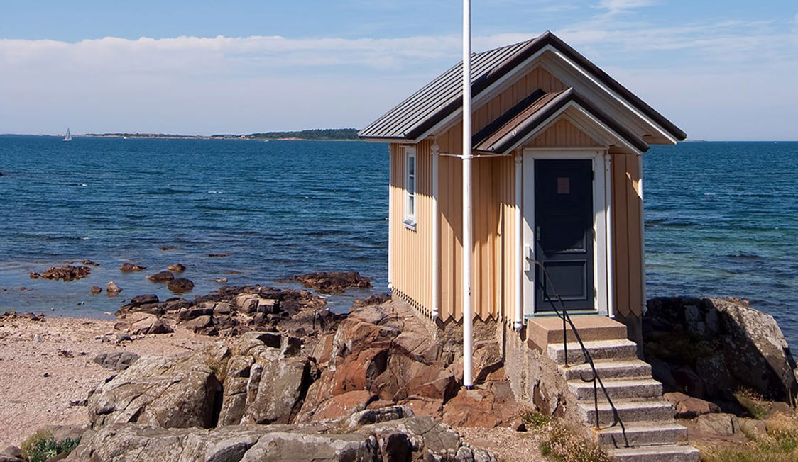 Lille træhus i gule nuancer stående på klipper ved havet under blå himmel.
