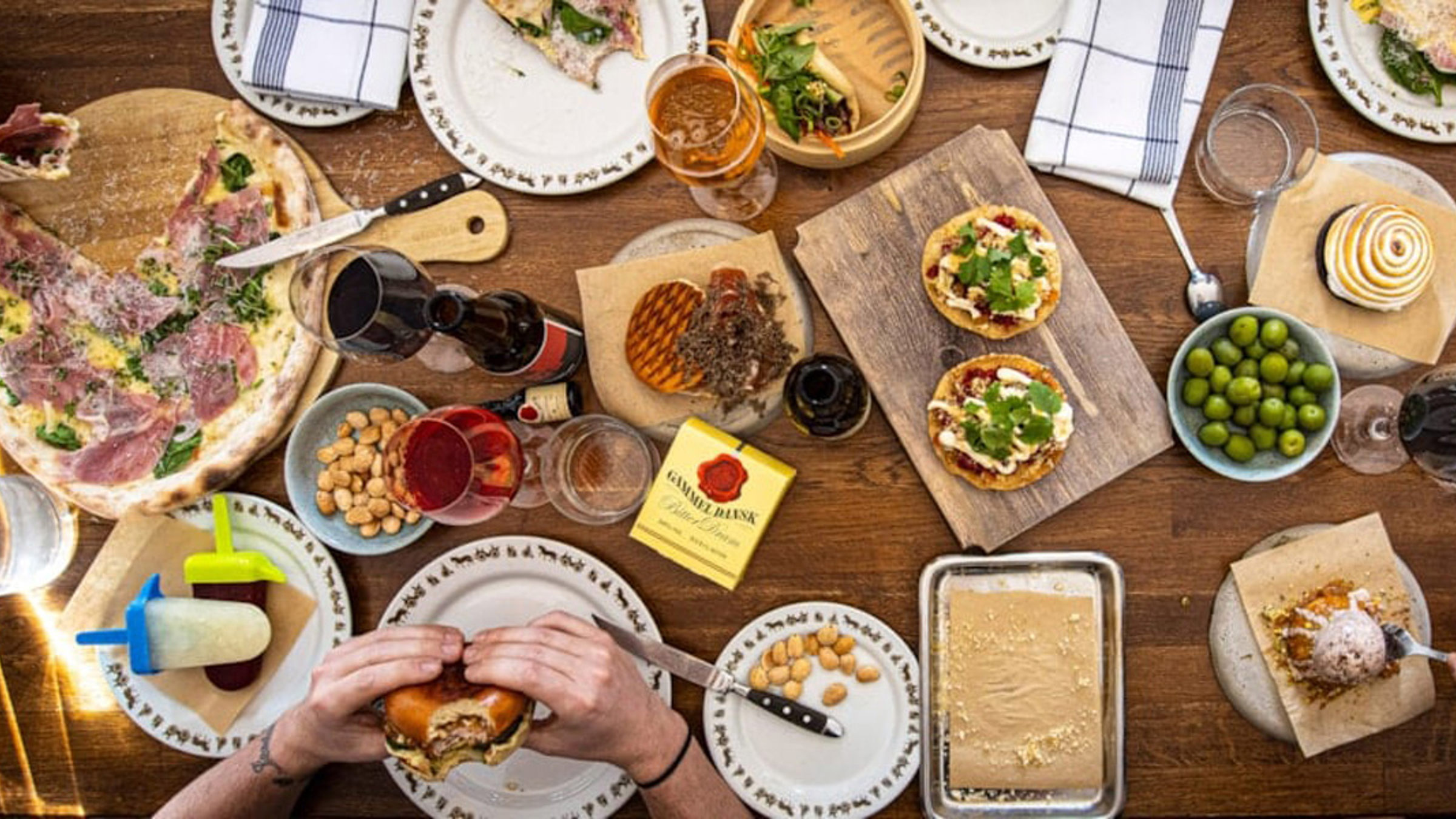 Mennesker nyder forskellige retter ved et træbord, herunder pizza, små tacos, oliven og drikkevarer.