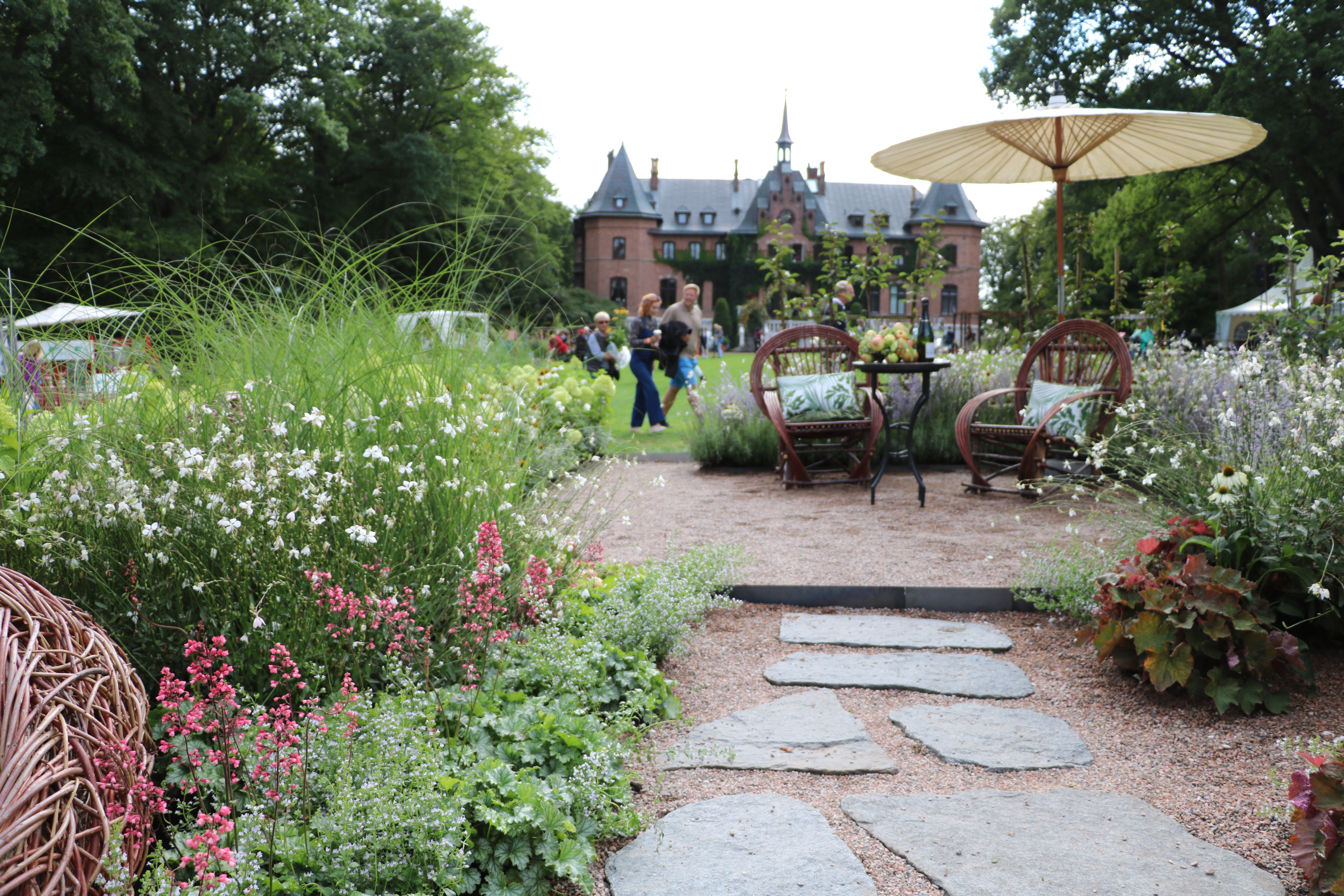 Stenbelagt sti fører til have med blomsterbede, kurvestole og lille bord under parasol i slotshave.