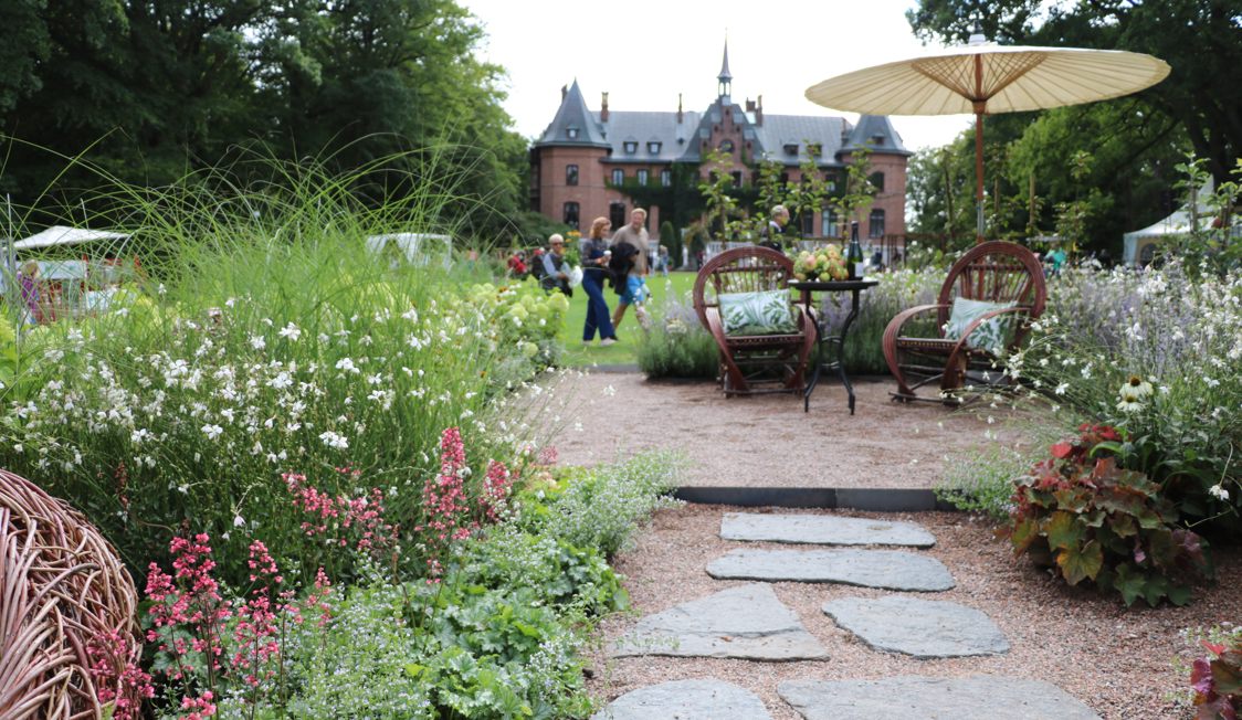 Stenbelagt sti fører til have med blomsterbede, kurvestole og lille bord under parasol i slotshave.