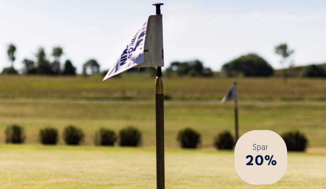 Golfbane med hvidt flag på pinden i forgrunden, grønne marker og træer i baggrunden.