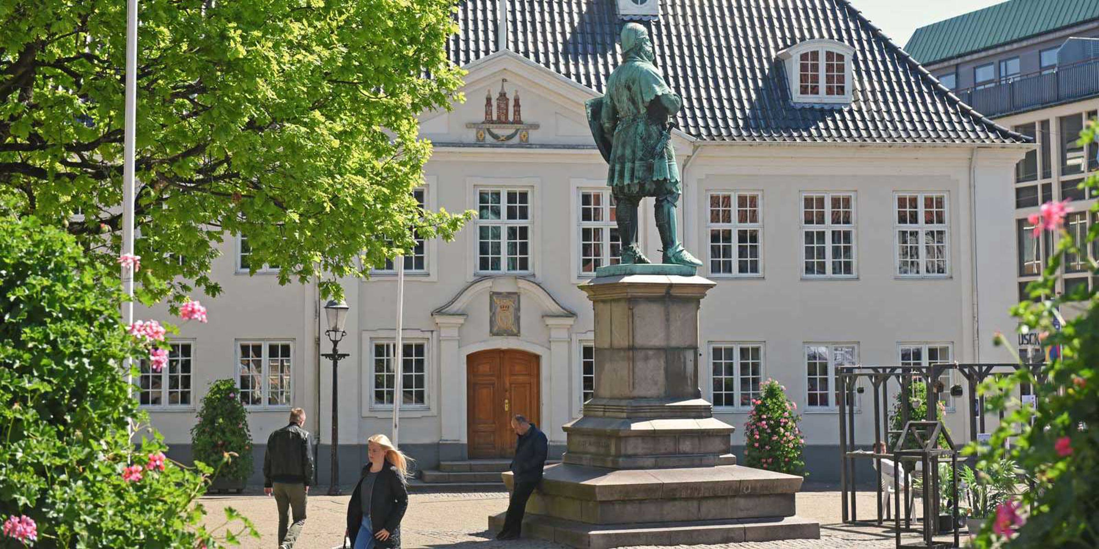 Statuen af en historisk figur står foran et gammelt, hvidt hus med klassisk arkitektur i solskin.