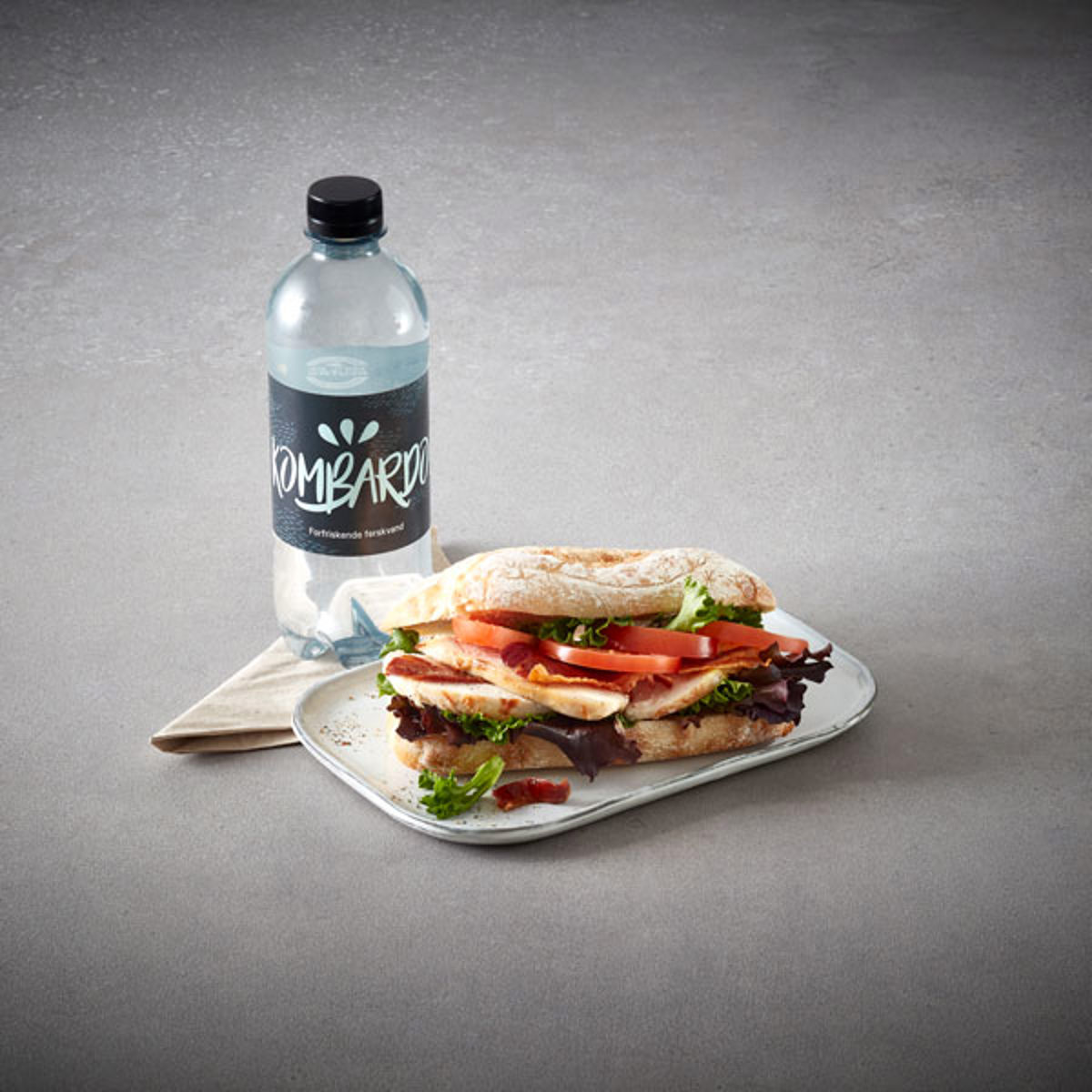 Kyllingesandwich med salat og tomater serveret på tallerken ved siden af flaske vand på gråt bord.