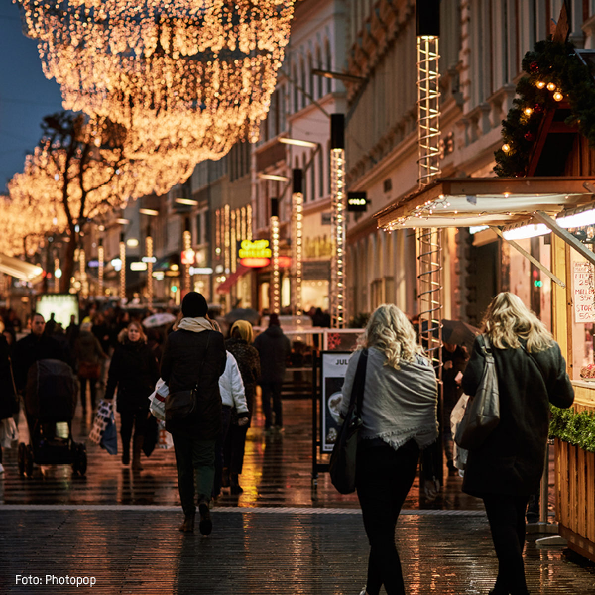 Gågade med julebelysning i en travl by, folk går og nyder aftenstemningen og dekorationerne.