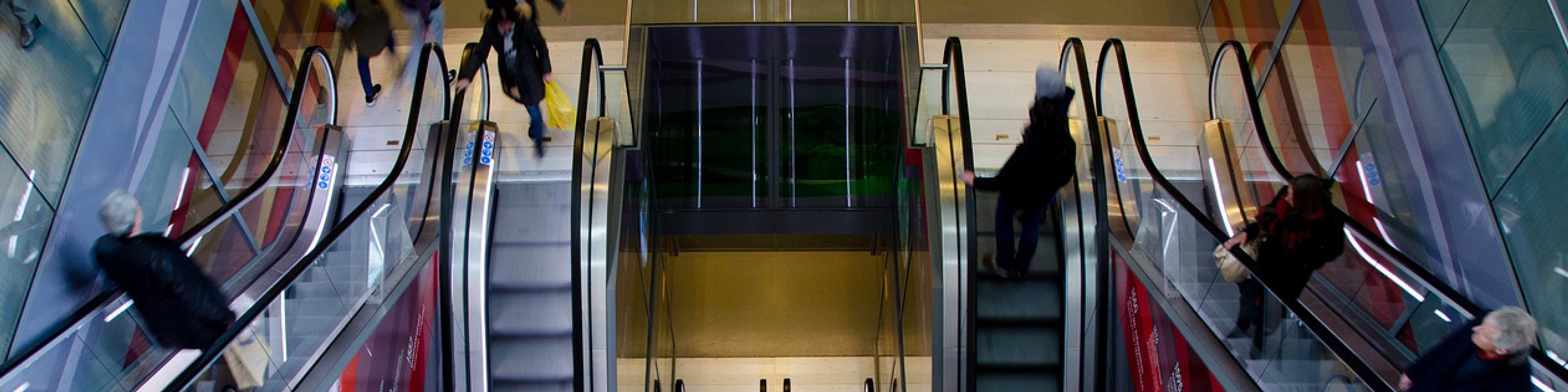 Personer bevæger sig op og ned ad rulletrapper i et travlt indkøbscenter fra et fugleperspektiv.