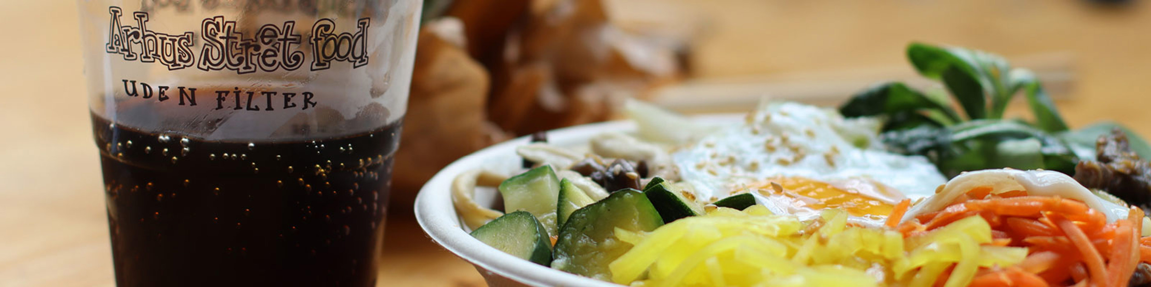 Skål med farverig asiatisk mad toppet med spejlæg ved siden af et glas sodavand fra Aarhus Street Food.