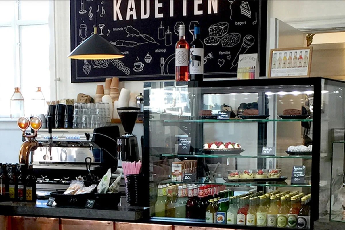 Caféinteriör med Kadetten Jpeg-skylt, kaffebryggare och konditorivaror i en modern miljö.