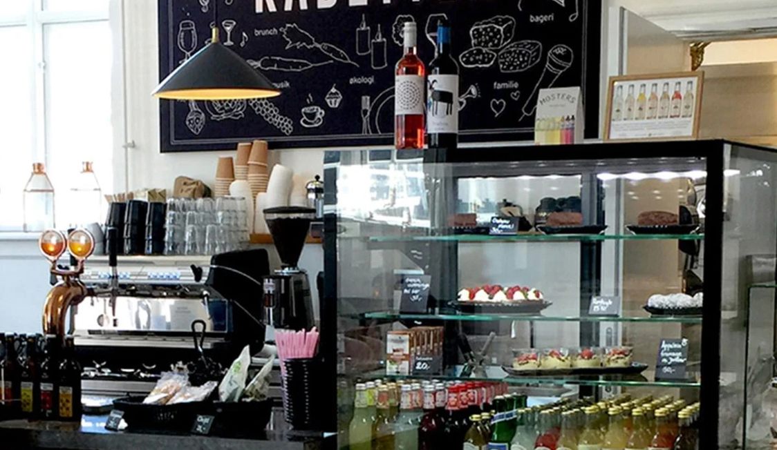 Caféinteriör med Kadetten Jpeg-skylt, kaffebryggare och konditorivaror i en modern miljö.