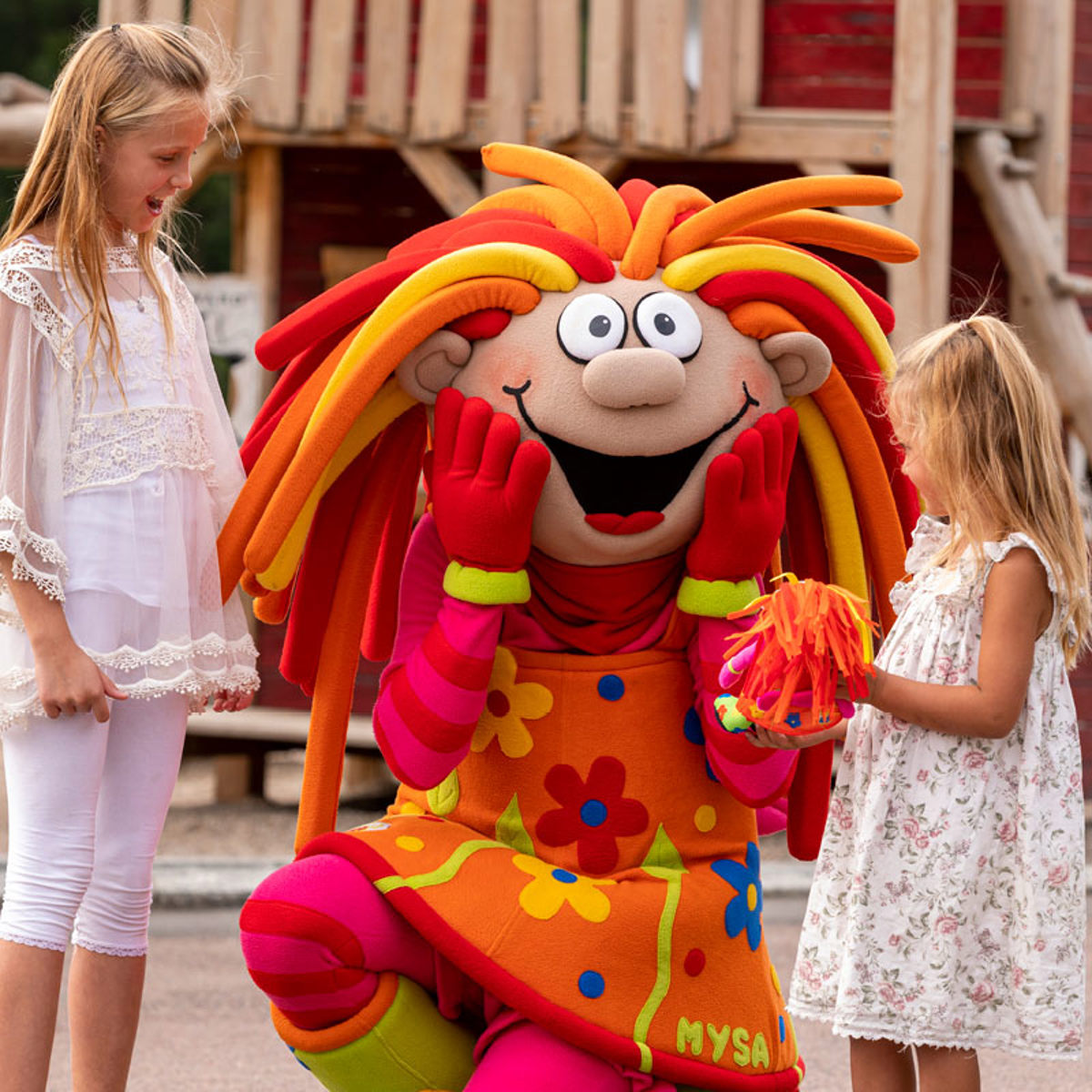 Farverig maskot i blomsterkjole underholder to glade piger i en sommerforlystelsespark.