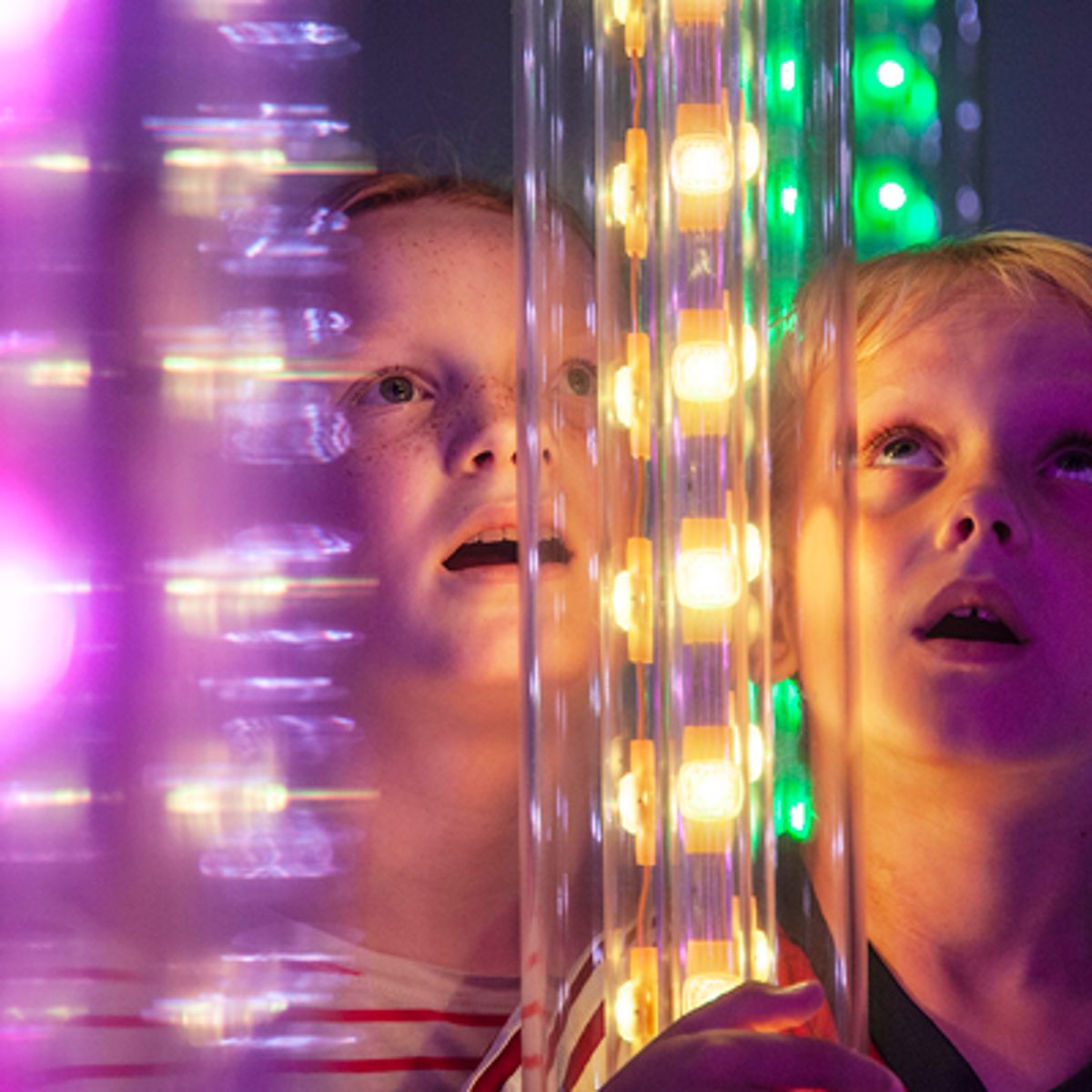 To børn betragter fascineret en lysinstallation med farverige, glødende pærer i en interaktiv udstilling.