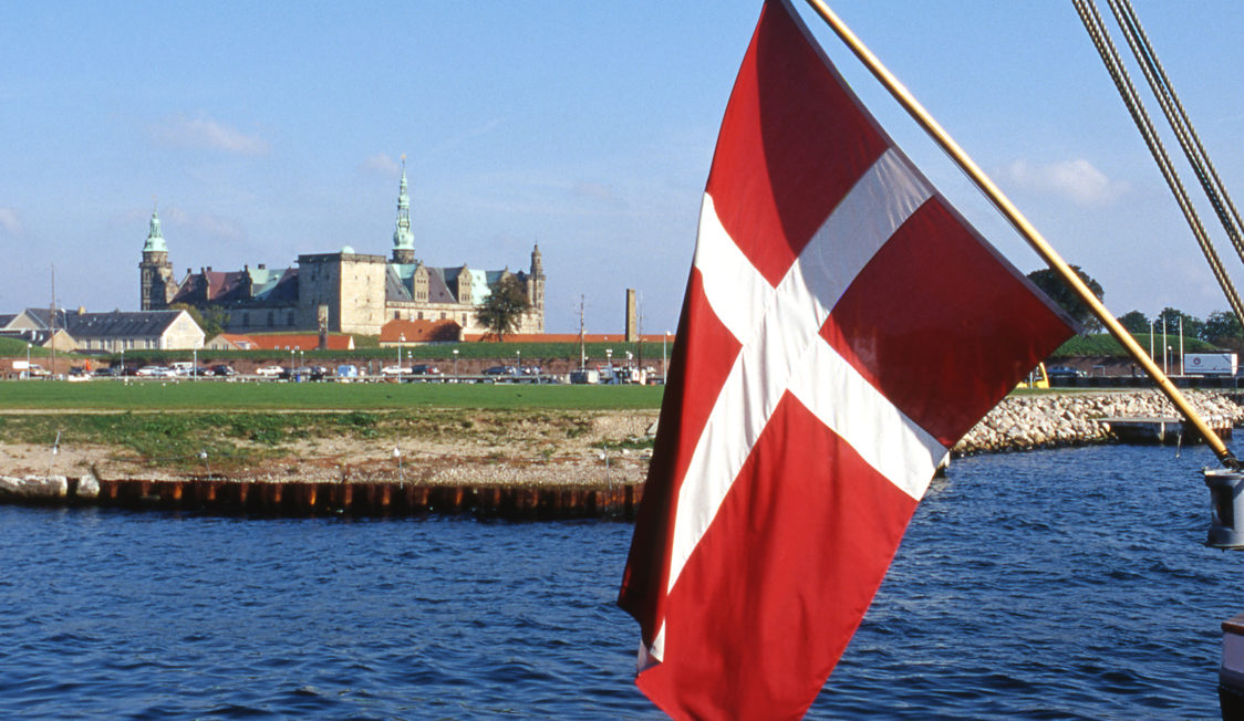 Dansk flagga vajar i förgrunden med utsikt över det historiska slottet vid kusten.