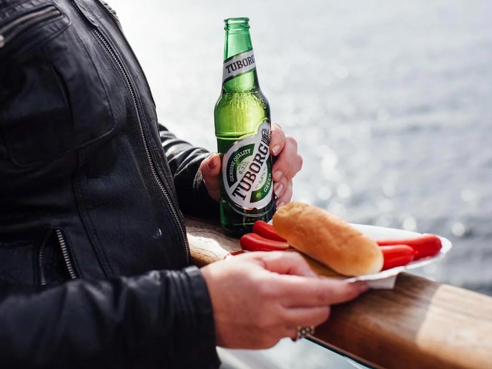 Person i læderjakke holder en Tuborg flaske og en rød pølse i brød ved vandoverfladen.