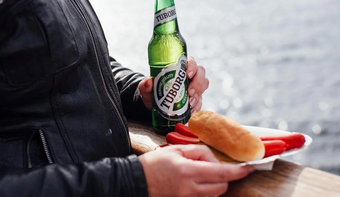 Person i læderjakke holder en Tuborg flaske og en rød pølse i brød ved vandoverfladen.