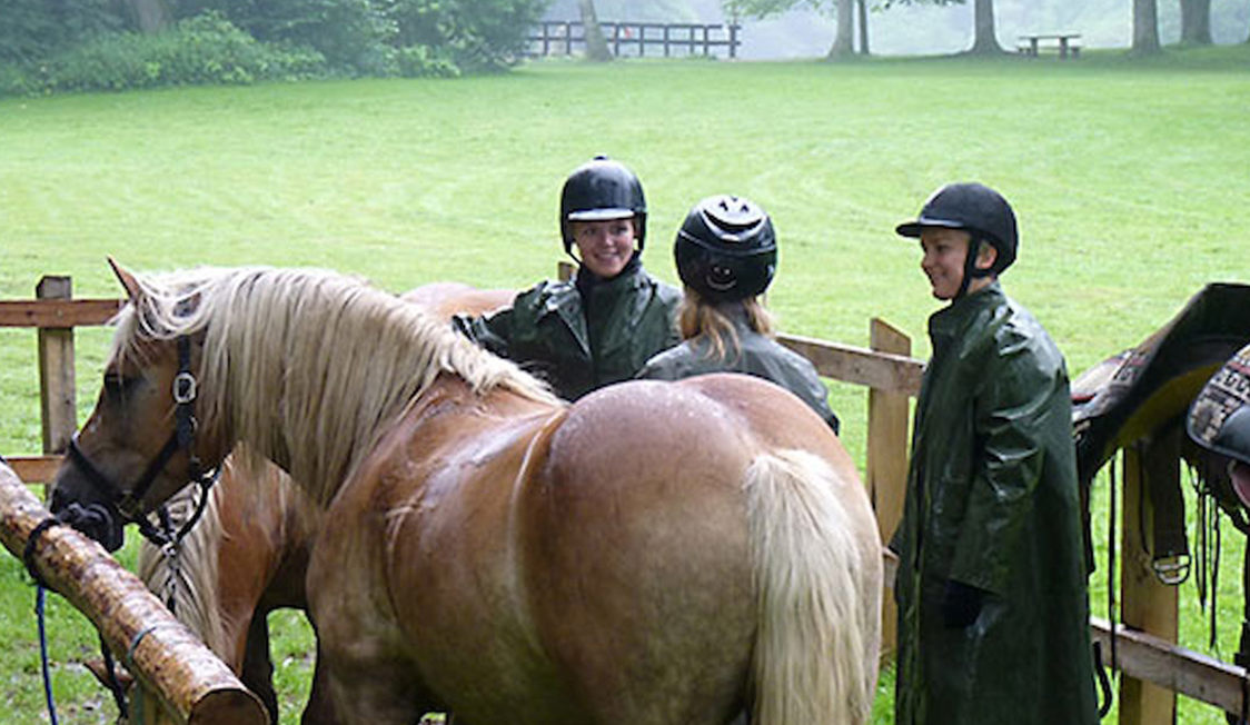 Ryttergruppe taler sammen ved hest på grøn græsplæne iført sikkerhedshjelme og regntøj.