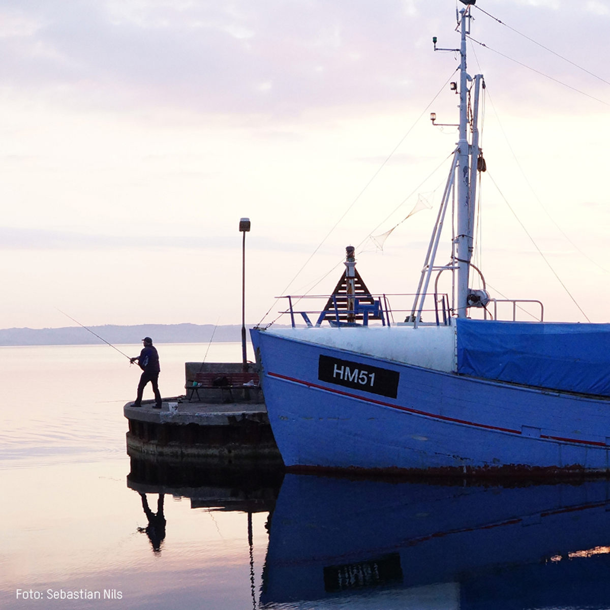 Fisker står på kajen og fisker ved siden af en blå fiskerbåd med navnet HM51 i stille vand ved solnedgang.