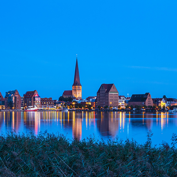 Vy över Rostocks stadssilhuett i skymningen, med vackra reflektioner i vattnet och klar blå himmel.