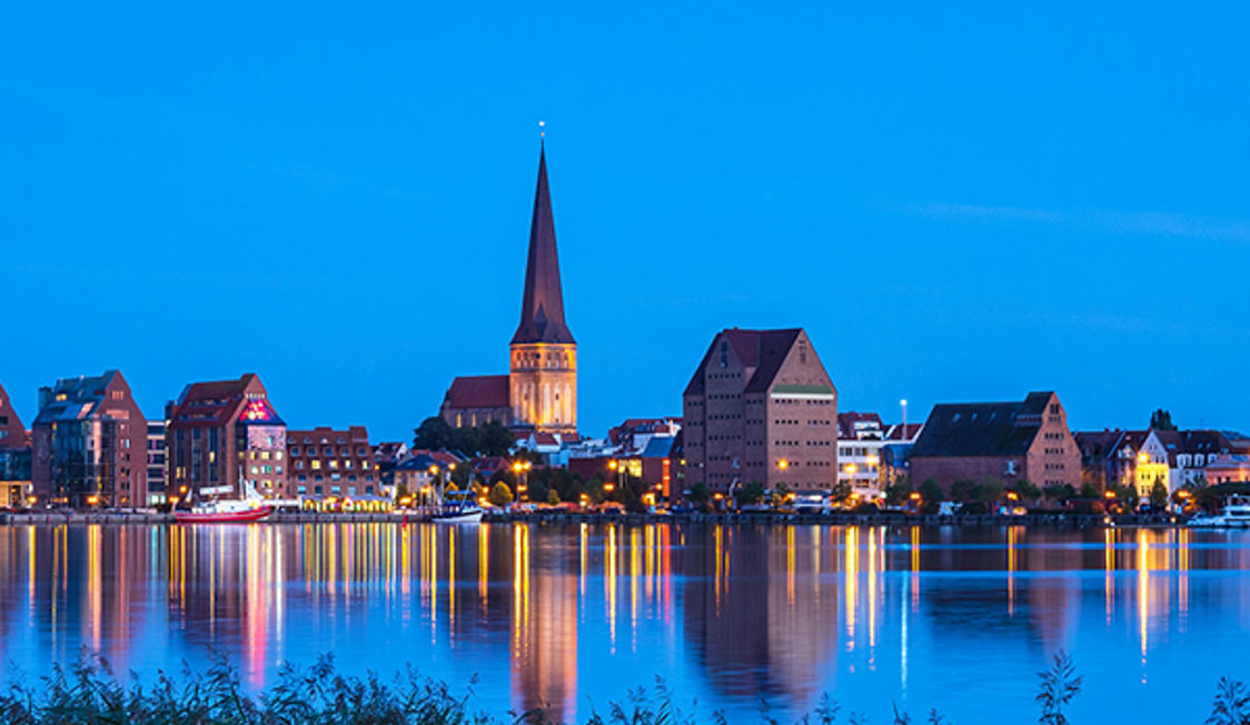 Vy över Rostocks stadssilhuett i skymningen, med vackra reflektioner i vattnet och klar blå himmel.